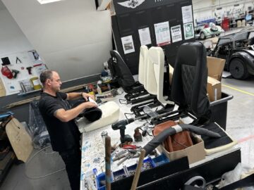 Een man werkt aan het monteren van autostoelen op een werkplek in een autowerkplaats. Op de tafel zijn verschillende gereedschappen, onderdelen en gedeeltelijk voltooide stoelen zichtbaar, die doen denken aan het nauwgezette vakmanschap dat te zien is bij Morgan Motor Company.