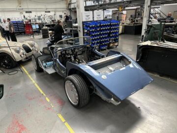 Een onvoltooide blauwe auto wordt gemonteerd in een autowerkplaats. Op de achtergrond zijn meerdere voertuigen en onderdelen zichtbaar, samen met werkplaatspersoneel. Tijdens de rondleiding kunt u uit de eerste hand getuige zijn van het nauwgezette vakmanschap dat een klassiek tintje belichaamt dat doet denken aan de nalatenschap van Morgan Motor Company.