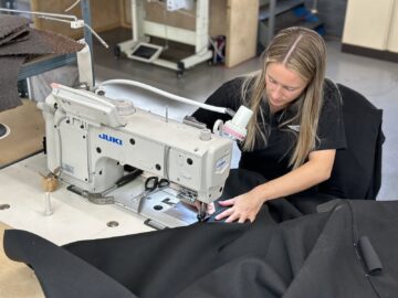 Een vrouw bedient een industriële Juki-naaimachine en naait in een werkplaats zwarte stoffen alsof ze een nauwgezette rondleiding bij de Morgan Motor Company volgt. Op de achtergrond zijn verschillende stoffen onderdelen en uitrustingen zichtbaar, vergelijkbaar met de gedetailleerde montage van klassieke auto’s.