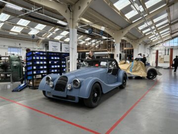 Twee klassieke auto's staan geparkeerd in een industriële garage. Eén auto is volledig zichtbaar en blauw geverfd, terwijl de andere gedeeltelijk bedekt is met beschermfolie. Op de achtergrond zijn mensen bezig, misschien ter voorbereiding op een rondleiding bij een vestiging als Morgan Motor Company.