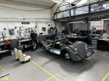 Een werkplaatsscène met een gedeeltelijk geassembleerde klassieke auto met gereedschap en uitrusting op omliggende banken. Een persoon werkt op een bankje, terwijl een ander op een bovenverdieping te zien is, misschien onderdeel van een rondleiding bij de gerenommeerde Morgan Motor Company.