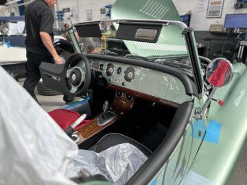 Een groene cabriolet, mogelijk een strak model van de Morgan Motor Company, wordt tentoongesteld in een garage met open dak en motorkap. Het interieur beschikt over een klassiek houten dashboard, terwijl er vlakbij een persoon in het zwart staat die een gedetailleerde rondleiding geeft over de kenmerken van de auto.