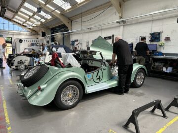 Monteurs werken aan een lichtgroene auto in vintagestijl in een goed verlichte garage. De motorkap van de auto is open en onthult de ingewikkelde onderdelen, terwijl op de achtergrond verschillende gereedschappen en uitrusting zichtbaar zijn. Het voelt als een kijkje in het nauwgezette vakmanschap van de auto's van Morgan Motor Company tijdens een exclusieve rondleiding.