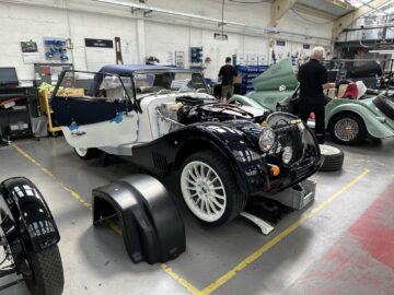 Een gedeeltelijk geassembleerde klassieke auto van Morgan Motor Company met open motorkap en deuren bevindt zich in een autowerkplaats. De motor is zichtbaar en er liggen verschillende gereedschappen en onderdelen verspreid. Op de achtergrond zijn verschillende medewerkers aanwezig die het ingewikkelde proces van het in elkaar zetten van deze iconische auto's laten zien aan de auto's tijdens een rondleiding.