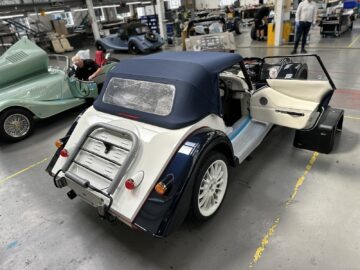 Achteraanzicht van een auto in vintage-stijl met blauwe en witte verf in een werkplaats, de deur open om het interieur te onthullen. Op de achtergrond verschijnen meerdere soortgelijke auto's en enkele mensen, die doen denken aan een scène uit de beroemde Morgan Motor Company tijdens een van hun informatieve rondleidingen.