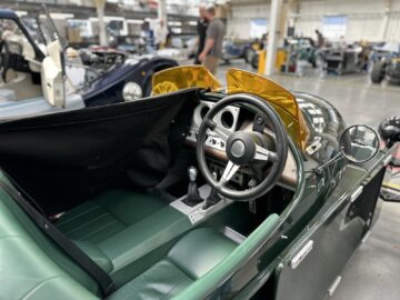 In een werkplaatsomgeving wordt een auto met open dak tentoongesteld, met gele windgeleiders en groenleren stoelen, een verbluffende creatie van Morgan Motor Company.