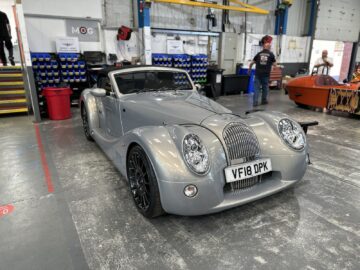 Een grijze vintage-stijl converteerbare sportwagen, waarschijnlijk een klassieker van Morgan Motor Company, staat geparkeerd in een garage met mensen en auto-uitrusting op de achtergrond. Op het kenteken staat "VF18 DPK.