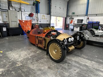 In een drukke werkplaats toont een op maat gemaakte driewielerauto van Morgan Motor Company met open motorkap ingewikkelde rode en gouden details. De auto staat omringd door diverse gereedschappen en apparatuur, wachtend op de volgende rondleiding.