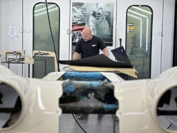 Een persoon die in een autolakwerkplaats werkt en zich concentreert op een stuk materiaal op een werkstation met auto-onderdelen op de voorgrond, wat doet denken aan het zelden auto's maken tijdens een rondleiding door Morgan Motor Company.