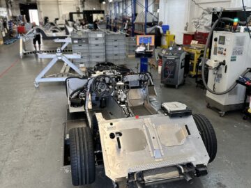 Het chassis van een gedeeltelijk geassembleerde auto staat op de vloer van een goed verlichte autowerkplaats, omringd door verschillende gereedschappen, apparatuur en machines. Het tafereel doet denken aan een Rondleiding bij de Morgan Motor Company.