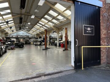 Weergave van het interieur van een autowerkplaats met meerdere auto's en een bord met het opschrift "Final Finish" op de open zwarte deur. De ruimte heeft een hoog plafond met meerdere lampen en zichtbare werkplekken, wat doet denken aan een Rondleiding Morgan Motor Company tour.