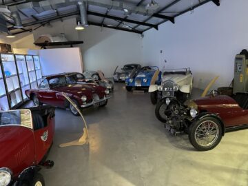 Een verzameling klassieke auto's in een museumomgeving, tentoongesteld in een goed verlichte, moderne ruimte. De vintage voertuigen, die doen denken aan auto’s gemaakt door ambachtslieden, variëren in kleur en stijl, waarbij de motorkappen en deuren openstaan om tentoongesteld te worden. Een Rondleiding Morgan Motor Company-tour versterkt de ervaring.