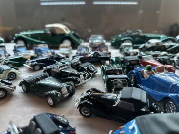 Een verzameling oldtimermodellen tentoongesteld op een oppervlak, met verschillende merken en kleuren, voornamelijk groen en zwart, met een onscherpe achtergrond. Deze tentoonstelling zou gemakkelijk deel kunnen uitmaken van een rondleiding bij de Morgan Motor Company, waarbij hun geschiedenis op het gebied van auto's wordt getoond.