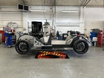 Een onafgewerkt autochassis van de Morgan Motor Company wordt in een werkplaats op een hydraulisch platform getild, waar het frame en de wielconstructies worden tentoongesteld tijdens de autorondleiding.