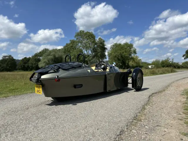 Une Morgan Super 3, avec sa teinte verte emblématique, est garée sur une route de campagne par une journée ensoleillée, avec des arbres et un ciel bleu avec des nuages en arrière-plan.