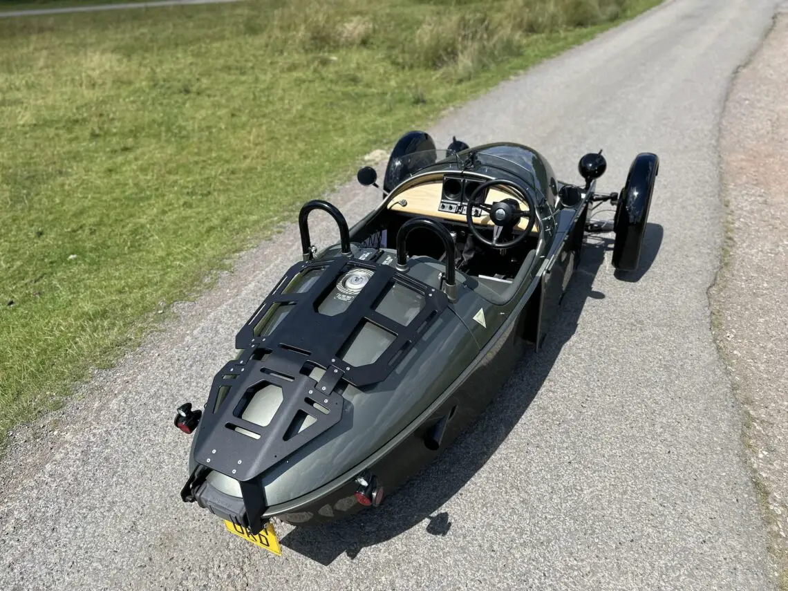 Een Morgan Super 3, met zijn gestroomlijnde ontwerp en open cockpit, staat geparkeerd op een landelijke weg nabij een grasveld.