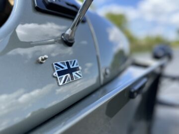 Gros plan de la carrosserie d'une Morgan Super 3 avec un petit emblème de l'Union Jack sur un extérieur gris, sous un ciel clair et partiellement nuageux.