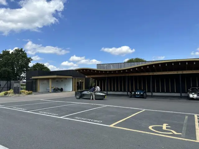 Een modern gebouw met grote houten overstekken grenst aan een parkeerplaats met gereserveerde plaatsen en één toegankelijke ruimte op de voorgrond. Tussen de oldtimers voor het gebouw valt een Morgan Super 3 op.