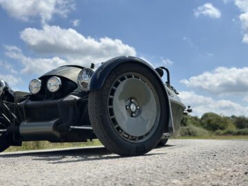 Tiefblick auf einen klassischen Morgan Super 3, mit Fokus auf das Vorderrad und die Scheinwerfer, geparkt auf einer asphaltierten Straße unter blauem Himmel mit vereinzelten Wolken.