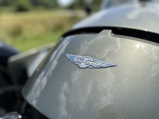 Vista frontal del capó de un coche Morgan Super 3, que muestra el emblema de la empresa con alas. El fondo muestra una vista borrosa de campos y árboles.