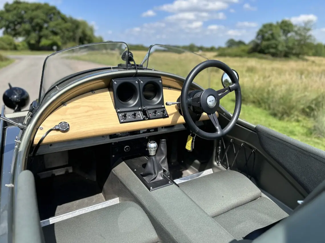 Interieur van een Morgan Super 3 cabriolet met een houten dashboard, zwart stuur en grijze stoelen. De auto staat geparkeerd op een landelijke weg met groene velden en bomen op de achtergrond.