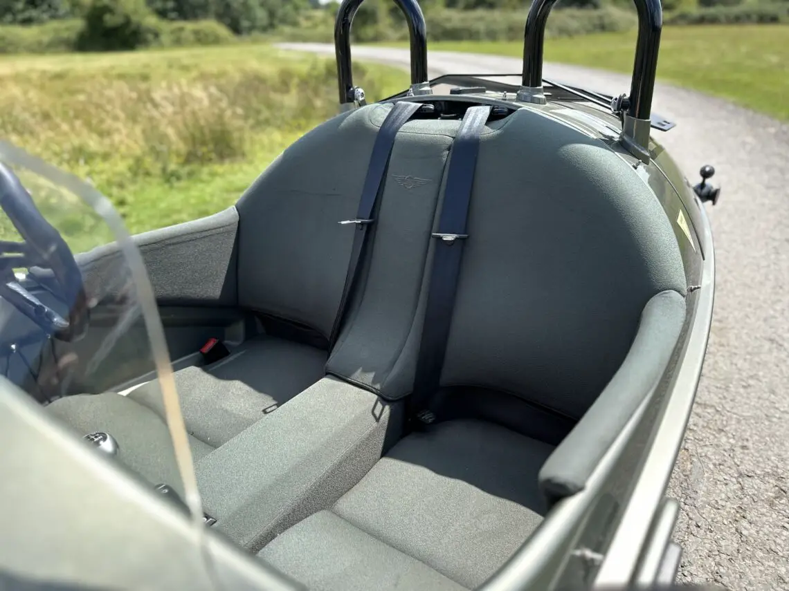 Un coche biplaza Morgan Super 3 gris con cinturones de seguridad negros, aparcado en una carretera cerca de campos verdes. La imagen se centra en el interior de los asientos y el parabrisas.