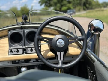 Close-up van het stuur en het dashboard van een klassieke Morgan Super 3-auto, met een zijspiegel die een schilderachtige buitenomgeving weerspiegelt.