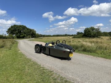 Ein dreirädriger Morgan Super 3 im Vintage-Stil fährt auf einer schmalen, kurvenreichen Straße, umgeben von grünen Feldern und unter einem teilweise bewölkten Himmel.