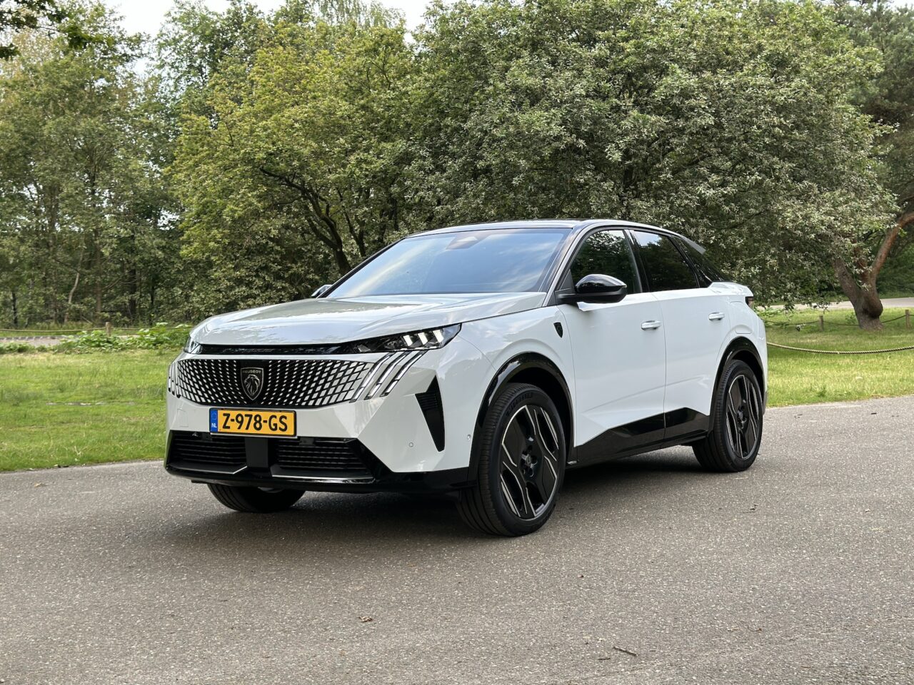 Een wit-zwarte Peugeot E-3008 SUV staat geparkeerd op een verharde weg met bomen en gras op de achtergrond. De auto heeft Nederlands kenteken (Z-978-GS).