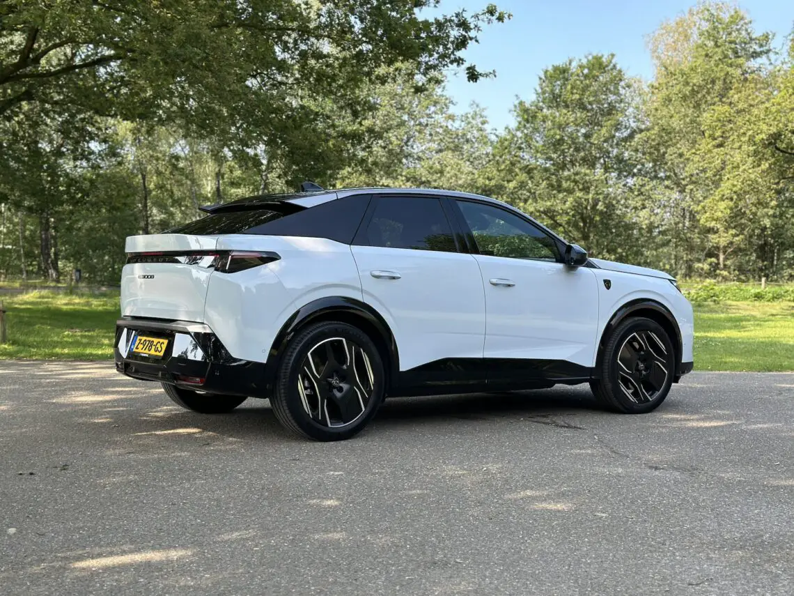 Een witte SUV Peugeot E-3008 staat geparkeerd op een verharde weg in een bosrijke omgeving, met bomen en groen op de achtergrond op een zonnige dag.