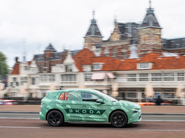 A Skoda car with camouflage trim drives past traditional buildings. The car is driving, with the background details somewhat blurred.