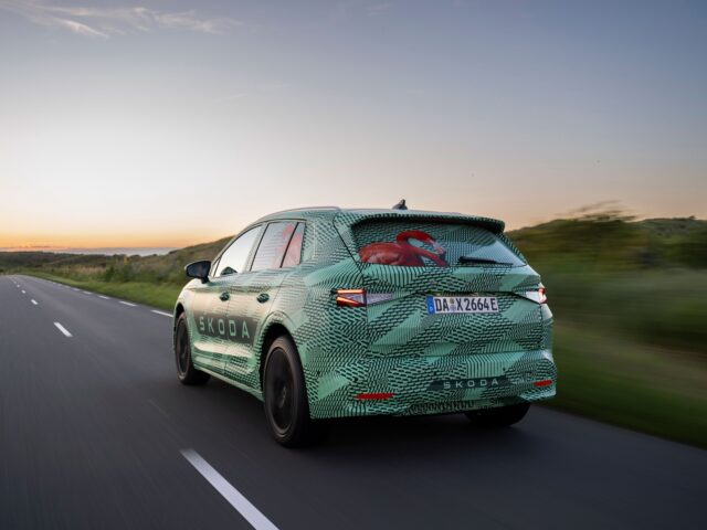A green Škoda car with camouflage pattern drives down a road during sunset, showing its rear and license plate number.