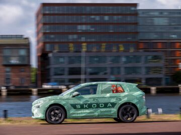 A green Skoda car drives along a waterfront road, with modern multi-story buildings in the background.