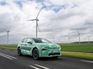 A camouflaged Skoda car driving on a road with spinning wind turbines in the background under a cloudy sky.