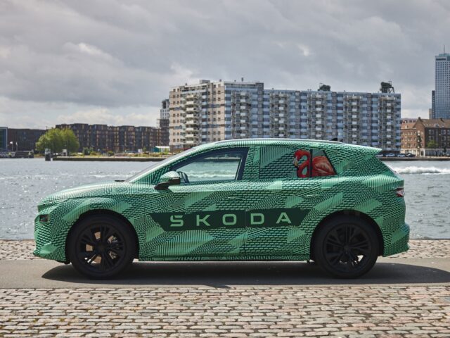 Een Skoda-auto met een groen en zwart camouflagepatroon staat geparkeerd vlakbij een waterkant met gebouwen met meerdere verdiepingen en water op de achtergrond.