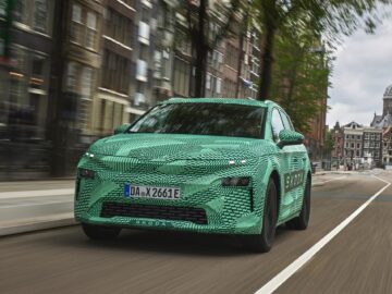 A green camouflaged Skoda car drives down a city street with vague buildings and a church in the background.