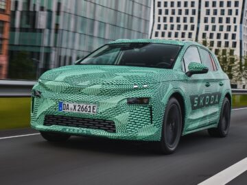 A camouflaged Skoda electric car drives down a city street with blurred buildings in the background.