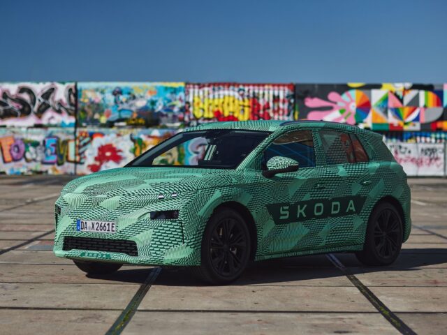 A Skoda car with a green-patterned exterior is parked in front of a wall covered in colorful graffiti on a bright day.