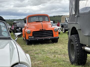 Een oranje vintage GMC-truck staat geparkeerd op een grasveld tussen andere klassieke auto's tijdens de West Oxon Steam & Vintage Show 2024, met verschillende mensen zichtbaar op de achtergrond onder een bewolkte hemel.