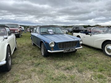 Een klassieke blauwe auto met het kenteken "HGT 223K" wordt tentoongesteld op een grasveld tussen andere vintage voertuigen onder een bewolkte hemel tijdens de West Oxon Steam & Vintage Show 2024.