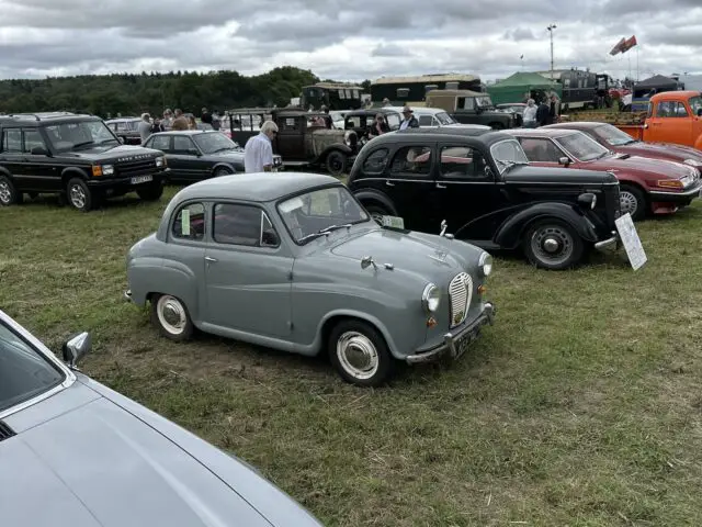 Een rij oldtimers buiten tentoongesteld op een grasveld onder een bewolkte hemel, met lopende mensen en andere voertuigen op de achtergrond, vastgelegd tijdens de West Oxon Steam & Vintage Show 2024 voor een fotoverslag.