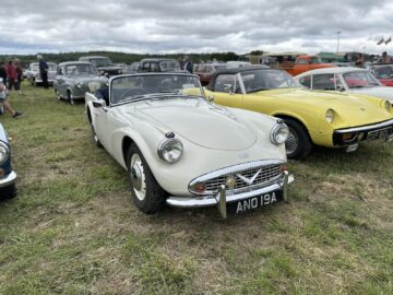 Tijdens de West Oxon Steam & Vintage Show 2024 wordt een reeks klassieke auto's buiten op een grasveld tentoongesteld, met een witte cabriolet op de voorgrond en diverse andere vintage voertuigen zichtbaar op de achtergrond, perfect voor een fotoverslag.