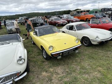 Een gele cabrioletsportwagen staat geparkeerd tussen andere klassieke voertuigen op de West Oxon Steam & Vintage Show 2024, terwijl mensen lopen en de auto's bekijken onder een bewolkte hemel. FOTOVERSLAG legt dit levendige moment vast.