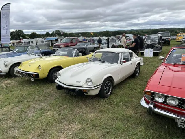 Een verzameling klassieke auto's, waaronder gele, witte en rode modellen, wordt buiten tentoongesteld op de Vintage Show 2024 in West Oxon Steam, waarbij bezoekers de voertuigen onder een bewolkte hemel onderzoeken.