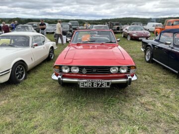 Een rode klassieke Triumph-auto met kenteken EMR 673L, tentoongesteld op de West Oxon Steam & Vintage Show 2024, omringd door andere vintage voertuigen, werd prachtig vastgelegd in dit Fotoverslag.