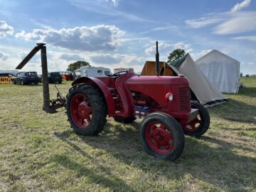 Een rode vintage tractor staat geparkeerd in een grasveld in de buurt van tenten en andere voertuigen op een zonnige dag tijdens de West Oxon Steam & Vintage Show 2024.