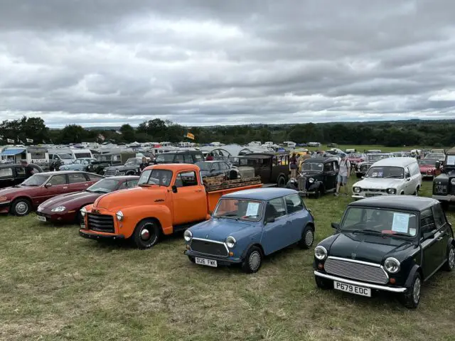 Tijdens de West Oxon Steam & Vintage Show 2024 worden verschillende klassieke auto's en vrachtwagens tentoongesteld op een buitenveld. De lucht is bewolkt en op de achtergrond zijn andere voertuigen en mensen zichtbaar.