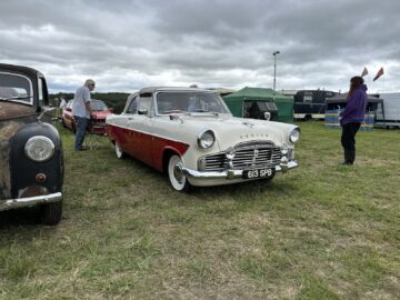 Een klassieke tweekleurige auto met het kenteken "613 SPB" staat geparkeerd op het gras tijdens de West Oxon Steam & Vintage Show 2024, omringd door andere vintage voertuigen, mensen en tenten.