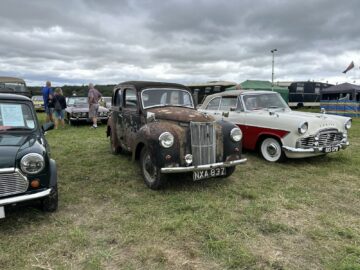 Een reeks oldtimers die buiten op het gras onder een bewolkte hemel worden tentoongesteld tijdens de West Oxon Steam & Vintage Show 2024. In het midden wordt een roestig voertuig geflankeerd door een kleine groene auto aan de linkerkant en een glimmende wit-rode auto aan de rechterkant, perfect voor elk fotoverslag.