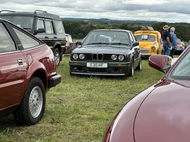 Een oldtimershow, onderdeel van de West Oxon Steam & Vintage Show 2024, toont verschillende klassieke auto's die op gras geparkeerd staan, waaronder een zwarte BMW E30, een rode sportwagen, een zwarte SUV en een gele auto met mensen die op de achtergrond slenteren. Kijk of de FOTOVERSLAG alle hoogtepunten vastlegt!